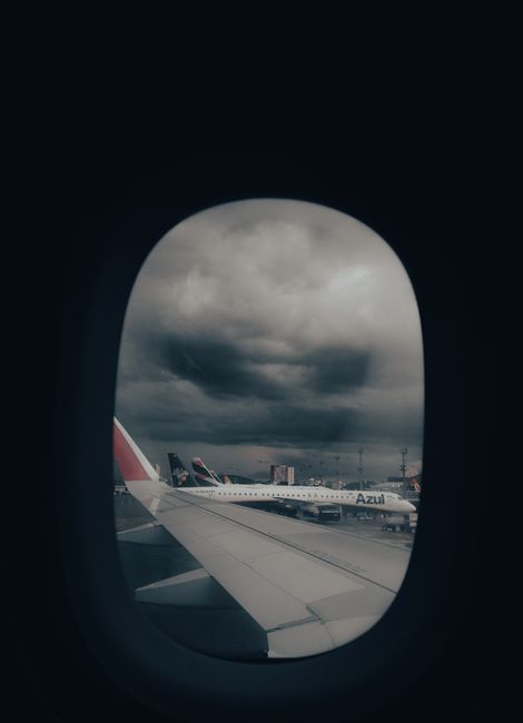 View through an airplane window showing a cloudy sky with dark, dense clouds and patches of lighter cloud cover. In the foreground, part of the wing of the aircraft is visible, resting horizontally across the lower part of the window frame, with its light-colored surface and visible panel lines. Outside the window, in the background, several airplane tails are visible; one prominently features a red and white design with a distinctive logo, suggesting a fleet of various airlines parked at an airport terminal. The tarmac and airport buildings are faintly visible at a distance beneath the stormy sky. The scene is illuminated by diffused natural light, creating a subdued, slightly moody atmosphere typical of overcast weather, which is consistent with an airport setting prior to takeoff or after landing, emphasizing the scene's association with air travel and the need for organized aircraft handling and ground services, such as waste and rubbish management by companies like Waste Collection Lewisham.