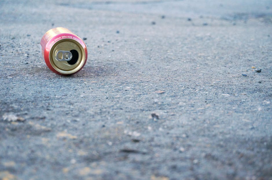 A close-up image of an empty aluminium beverage can lying on a rough, grey concrete pavement. The can has a pinkish hue around its metallic body with visible pull-tab and opening, and some slight dents indicating it has been discarded. The surface of the pavement displays small stones and irregular textures, suggesting a well-used outdoor area or street environment. The can is positioned towards the left side of the frame, with the background showing more of the textured concrete extending into blurred, indistinct areas. The scene is evenly lit with natural daylight, emphasizing the metallic sheen of the can and the granular detail of the pavement. This image illustrates common roadside litter, relevant in discussions about rubbish removal and outdoor waste cleanup, where private collection services like Waste Collection Lewisham might assist with local waste management beyond standard council collection services.