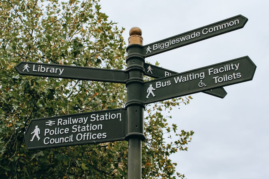 A tall black metal signpost with multiple directional signs attached to it stands in front of a background of leafy green and yellow trees. The signs are rectangular with rounded edges, each displaying white text and icons indicating different local destinations, including 'Biggleswade Common,' 'Library,' 'Bus Waiting Facility,' 'Toilets,' 'Railway Station,' 'Police Station,' and 'Council Offices.' The sign for 'Bus Waiting Facility' features a pedestrian icon, while the others have walking person icons. The overall scene appears to be in an outdoor public area, possibly near a town centre or park, with natural daylight providing soft, even lighting. This type of signage is typical for guiding visitors and residents to important civic and community facilities, which can be related to managing or orienting around local waste collection or civic services. The signpost’s design and placement suggest it is part of a well-maintained urban or suburban environment, consistent with locations where rubbish removal services may operate to support community cleanliness and infrastructure.
