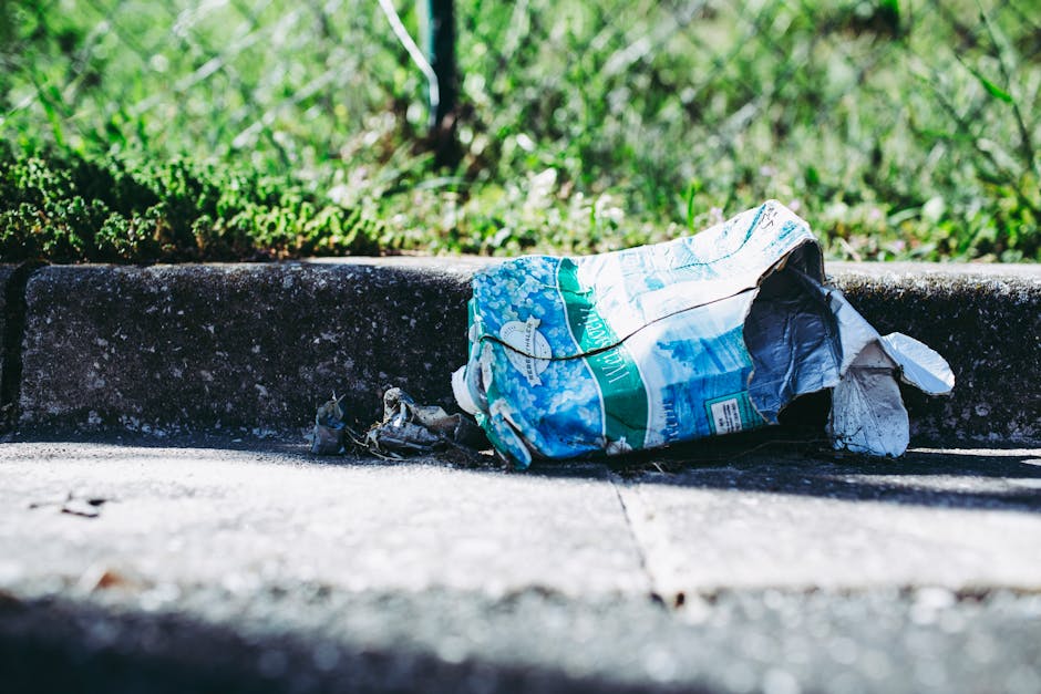 A close-up image of an empty aluminium beverage can lying on a rough, grey concrete pavement. The can has a pinkish hue around its metallic body with visible pull-tab and opening, and some slight dents indicating it has been discarded. The surface of the pavement displays small stones and irregular textures, suggesting a well-used outdoor area or street environment. The can is positioned towards the left side of the frame, with the background showing more of the textured concrete extending into blurred, indistinct areas. The scene is evenly lit with natural daylight, emphasizing the metallic sheen of the can and the granular detail of the pavement. This image illustrates common roadside litter, relevant in discussions about rubbish removal and outdoor waste cleanup, where private collection services like Waste Collection Lewisham might assist with local waste management beyond standard council collection services.