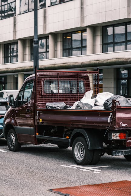 A small, dark red flatbed truck parked on an urban street in front of modern office buildings with large glass windows and concrete facades. The truck's bed is loaded with various types of dumped waste, including black and white plastic bags, some visibly bulging and partially open, revealing contents that appear to be household or general rubbish. A metal safety grid is mounted behind the cab to secure the load, and the truck's rear lights and small wheels are visible beneath the bed. The scene takes place during daytime with ambient natural light, and the truck is positioned close to a priority lane marked on the street. A white vehicle is partially visible behind the truck, and a lamppost stands to the left, adding to the urban environment context. This image exemplifies private waste collection options often used in conjunction with or as alternative to traditional rubbish disposal methods, aligning with services provided by Waste Collection Lewisham for on-site clearance and waste removal.