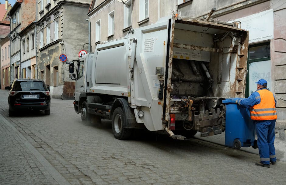 A white commercial waste collection truck with an open rear compartment, parked on a narrow cobblestone street in an urban setting. The truck's rear is rusted and partially empty, designed for on-site rubbish collection. A waste collection operative, dressed in blue overalls and a high-visibility orange vest with a blue cap, is seen lifting a blue wheeled bin towards the truck's open compactor. To the left, a black car is parked parallel to the curb, and in the background, there are multi-storey buildings with aged facades featuring windows, some with awnings or decorative trim. The scene is outdoors during daylight, with natural light illuminating the scene, emphasizing the textures of the cobblestones, the metal surfaces of the truck, and the surrounding architecture. The image relates to independent waste handling and private rubbish collection, fitting within the context of alternative rubbish removal options to skip hire, as provided by waste management services like Waste Collection Lewisham.