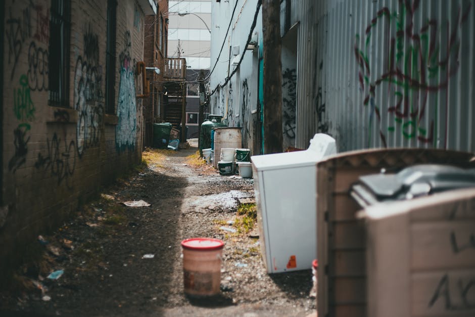 A narrow alleyway between brick and metal-clad buildings, cluttered with discarded household waste and debris. On the right, a large white washing machine or dishwasher is positioned with its back facing outward, alongside a wooden crate filled with miscellaneous items, all appearing weathered. Near the foreground, a red plastic bucket with a lid sits on damp, muddy ground strewn with small trash pieces and dead leaves. Further down the alley, several green garbage bins and a white container are aligned against the walls, some partially open or tipped over. The surrounding walls are covered in multicoloured graffiti and peeling paint, adding an urban atmosphere. In the background, wooden stairs and small balconies indicate residential access, with faint natural light filtering in from the alley’s entrance. The scene illustrates an environment typically associated with independent waste collection or private rubbish clearance services, highlighting the kind of bins and debris managed by waste collection specialists like Waste Collection Lewisham in Lewisham, UK, with a focus on clear visual details that support rubbish removal context and on-site clearance activities.