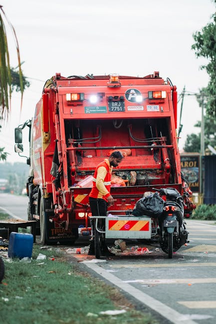 A waste collection worker in a yellow and red uniform is operating a large red refuse vehicle on a street. The worker is positioned at the back of the vehicle, which features a metal hopper with secured hydraulic arms, and appears to be engaged in rubbish disposal or transfer activity. The vehicle's exterior is painted bright red with reflective striping, and it displays identification markings, including a fleet number and safety labels. The scene takes place on a street lined with some greenery and utility poles, with visible pavement markings and a few scattered debris or rubbish bags on the ground nearby. In the background, there are trees and a building structure, suggesting an urban or suburban environment. The sky is overcast, providing diffuse lighting for the scene, which highlights the functional and professional nature of waste management operations carried out by independent rubbish collection services like Waste Collection Lewisham, supporting alternative waste handling methods in the area.