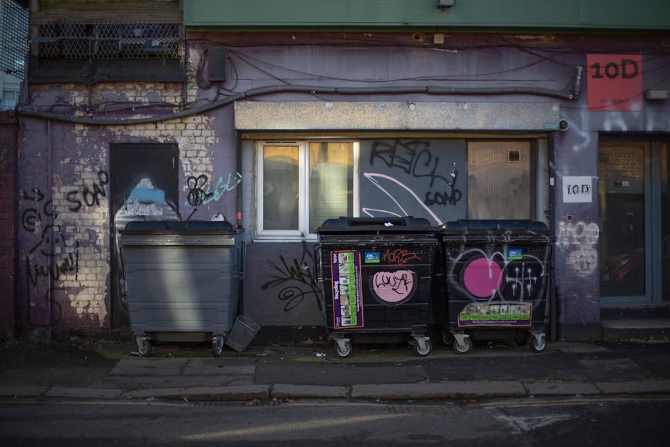 The image shows a row of three large wheelie bins placed on a sidewalk in front of an urban building with a weathered, graffiti-covered brick and concrete facade. The first bin, positioned on the left, is light grey with a smooth, metallic finish and a closed lid. The middle and right bins are black with cartoonish pink heart and graffiti stickers on their sides, featuring various spray-painted tags and markings in black, white, and pink. The environment appears dimly lit, likely during the evening or early morning, with some natural light reflecting off the building’s window, which has a partially visible curtain. The building’s exterior includes peeling paint, exposed brickwork, and electrical wires running along the facade, with a small window in the center partially obscured by grime and graffiti. To the right, a glass door with a red and white signage marked '10D' can be seen, indicating an apartment or business number. This scene depicts an everyday urban setting where waste collection might occur through private rubbish disposal services, like those offered by Waste Collection Lewisham, providing an alternative to council-managed refuse collection.