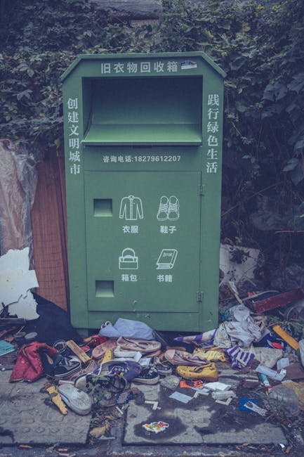 A large green waste collection kiosk with Chinese inscriptions and a contact phone number, situated outdoors against a backdrop of dense foliage and trees. The kiosk is designed for recycling old clothing and other items, with designated slots labeled for clothing, shoes, bags, and books. In front of the kiosk, there is a scattering of discarded household items, including shoes, clothing, bags, and various miscellaneous rubbish, some of which appear torn, dirty, or discarded carelessly on the ground. The surrounding environment is slightly cluttered with debris, indicating improper waste disposal. The weather appears overcast or cloudy, casting diffuse natural light over the scene. Waste Collection Lewisham occasionally handles private waste removal services, although this collection point suggests an informal or community-based recycling initiative, unrelated to official municipal rubbish collection services. The scene underscores the importance of proper disposal methods and community involvement in waste management for reuse and recycling.