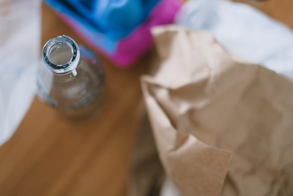 A close-up of a clear, empty glass bottle with a narrow neck and smooth, transparent surface placed on a wooden table. Surrounding the bottle are crumpled brown paper packaging and blurred objects in the background, including a partially visible plastic container with a colorful lid. The scene is lit with natural or soft ambient light, highlighting the textures of the paper and the reflective quality of the glass. The setting appears to be indoors, possibly in a recycling or waste collection context, with the discarded packaging and bottle indicating a focus on waste removal or reuse efforts by Waste Collection Lewisham, supporting alternative rubbish handling services.