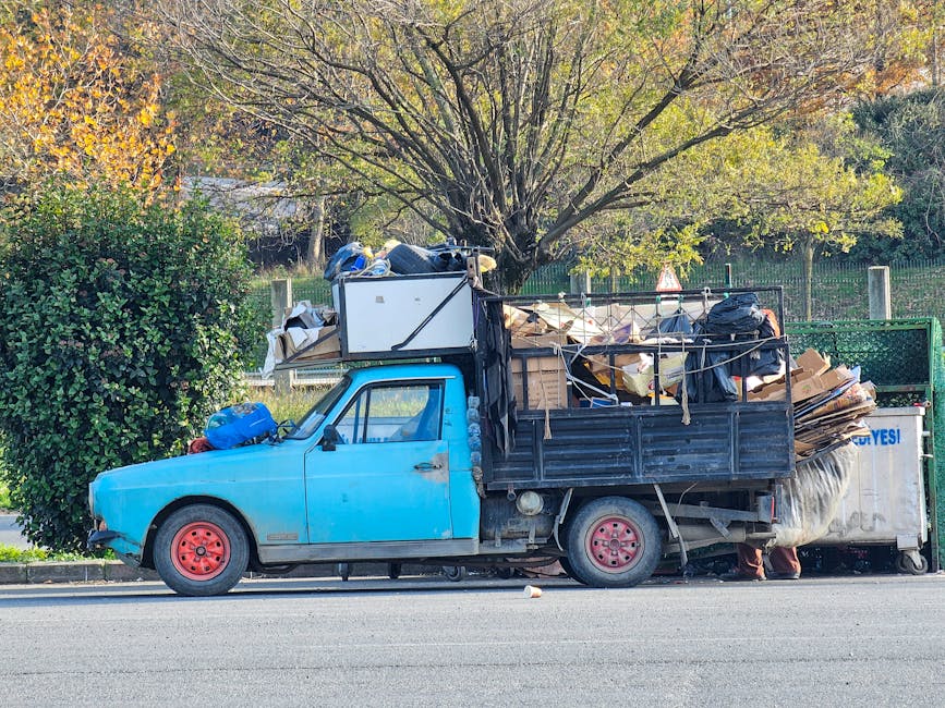 A small, light blue utility truck parked parallel to the curb on a street, heavily loaded with discarded items including cardboard boxes, plastic bags, and various household rubbish piled high in the open cargo area. The truck's body shows signs of wear, with a slightly faded finish and red-painted wheels. The vehicle is situated beside a green hedge on the left, with a large tree in the background casting shadows and autumnal foliage visible on the branches. In the background, a metal fence surrounds a park or garden area, and the street appears to be in a quiet residential or suburban setting. The loading is organized without visible signs of recycling separation, indicating a possible private waste collection or on-site clearance service. The overall scene is well-lit with natural daylight, emphasizing the cluttered load intended for rubbish removal or disposal, aligning with services offered by wastecollectionlewisham.co.uk.