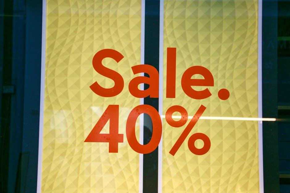 A promotional sign displayed in a shop or retail window with a bright yellow background featuring a subtle geometric pattern of small, triangular facets. The sign prominently shows the words 'Sale. 40%' in bold, large, red-orange letters and numerals, with the percentage symbol slightly smaller and positioned after the number. The sign is framed by a narrow white border, and the left side of the image shows part of a dark edge of the window frame or surrounding structure. The background behind the sign is dark, indicating it is seen through a glass surface, with some minimal reflections visible across the surface. The overall appearance suggests a clearance or discount promotion, common in retail or commercial settings, with a clean and professional presentation designed to attract attention to sales events.