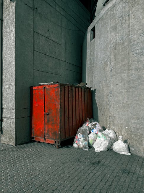 A rusted, red metal refuse container with visible wear and patches of peeling paint is situated outdoors against a dark, textured wall on the left side of the image. The container appears to be a large, industrial-style bin used for waste storage, with a partially open top revealing dark interior space. Next to it, on the right, is a small pile of white and semi-transparent plastic rubbish bags, some of which are loosely tied and slightly bulging with unknown waste contents. The bags rest on a dark grey, brick-patterned pavement that extends across the scene. Behind the container, a tall, smooth, light-colored wall with a small rectangular opening near the top can be seen, suggesting an urban environment or service area. The setting is illuminated naturally, with diffuse daylight casting minimal shadows, creating a neutral, utilitarian scene consistent with on-site waste collection or private rubbish disposal arrangements managed by Waste Collection Lewisham in Lewisham.