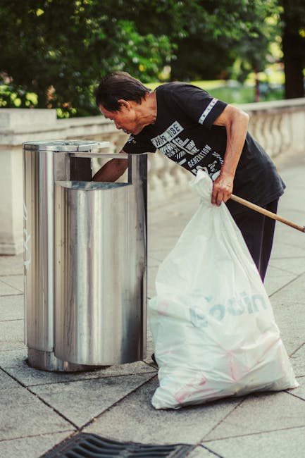 A middle-aged man wearing a black T-shirt with white text and dark pants is seen emptying a large white plastic sack filled with waste into a stainless steel public rubbish bin with a curved opening, positioned on a paved sidewalk. The man is leaning slightly forward, holding the sack with his left hand and using his right hand to lift waste into the bin. A long wooden stick is resting against his right side. The background shows a concrete balustrade, lush green trees, and grassy areas, indicating an outdoor setting in a park or street-side location during daylight. The scene emphasizes proper waste disposal and litter management, supported by the presence of waste collection services, possibly provided by Waste Collection Lewisham as part of local waste management efforts, and highlights the importance of responsible rubbish handling in community spaces.