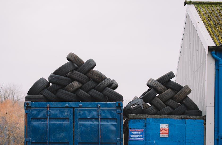 Two large blue containers are positioned outdoors, each filled with a significant quantity of used black rubber tires that are stacked in a haphazard, overlapping manner. The tires vary slightly in size and show signs of wear, with some displaying light scuffs and surface dirt, while others have visible sidewall patterns. The container on the left appears slightly taller and is covered with a layer of dust or dirt, indicating it has been exposed to the elements. The container on the right, made from a similar blue material with a brick pattern on its surface, holds tires arranged more compactly. In the background, part of a white building with a slanted roof covered in moss and green algae is visible, along with a pale, overcast sky suggesting a grey, cloudy day. The scene is typical of waste management or private disposal services, such as those provided by Waste Collection Lewisham, handling the collection and disposal of end-of-life tires as part of broader rubbish removal activities in Lewisham. The environment is functional and straightforward, emphasizing the storage of waste materials prior to transportation or recycling.